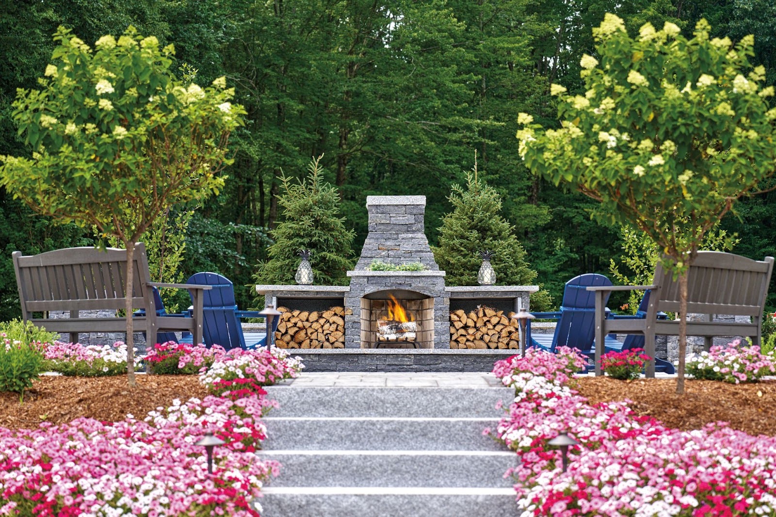 An outdoor living space featuring a stone fireplace, deck chairs and a stone patio surrounded by landscaped flower beds.