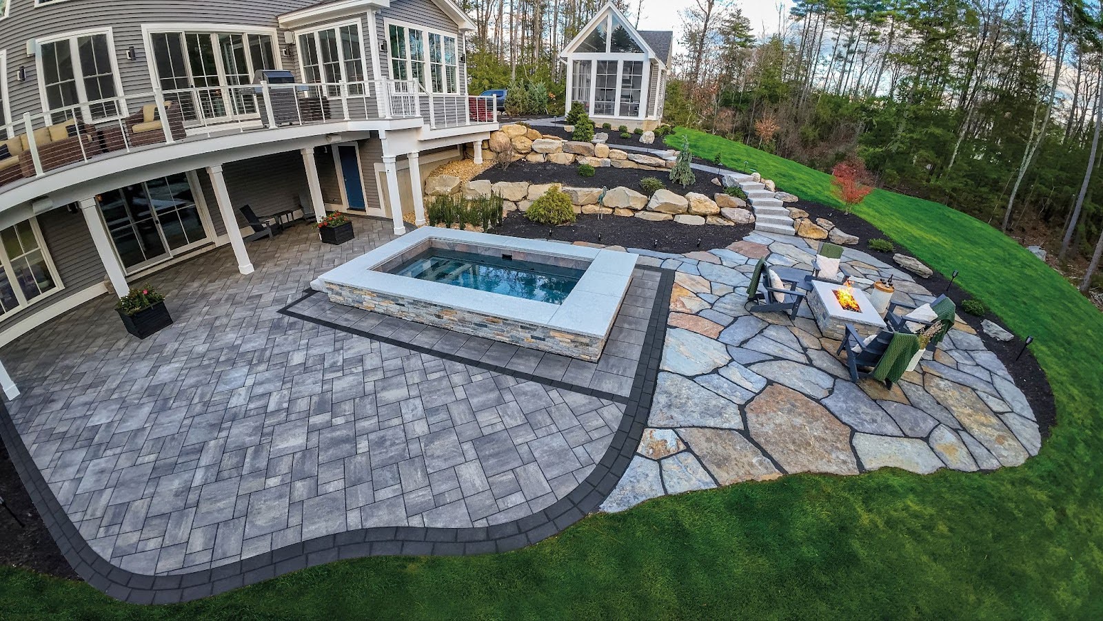 An outdoor hardscape retreat featuring a hot tub, fire pit and wicker seating arranged on a natural stone patio.